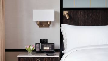 Ein Hotelzimmer mit einem Bett, einem Nachttisch mit Lampe, einem Thermostat und einer Vase mit Blumen.