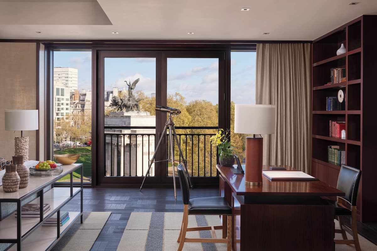 A modern room with a desk, chairs, a bookshelf, and a balcony with a view of buildings and a statue.