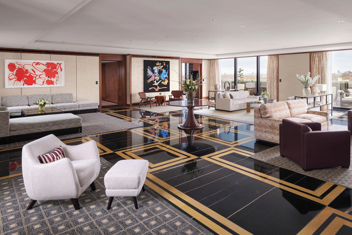 A spacious living area with a large sofa set, a coffee table, and a patterned rug. There is a large window offering a city view.