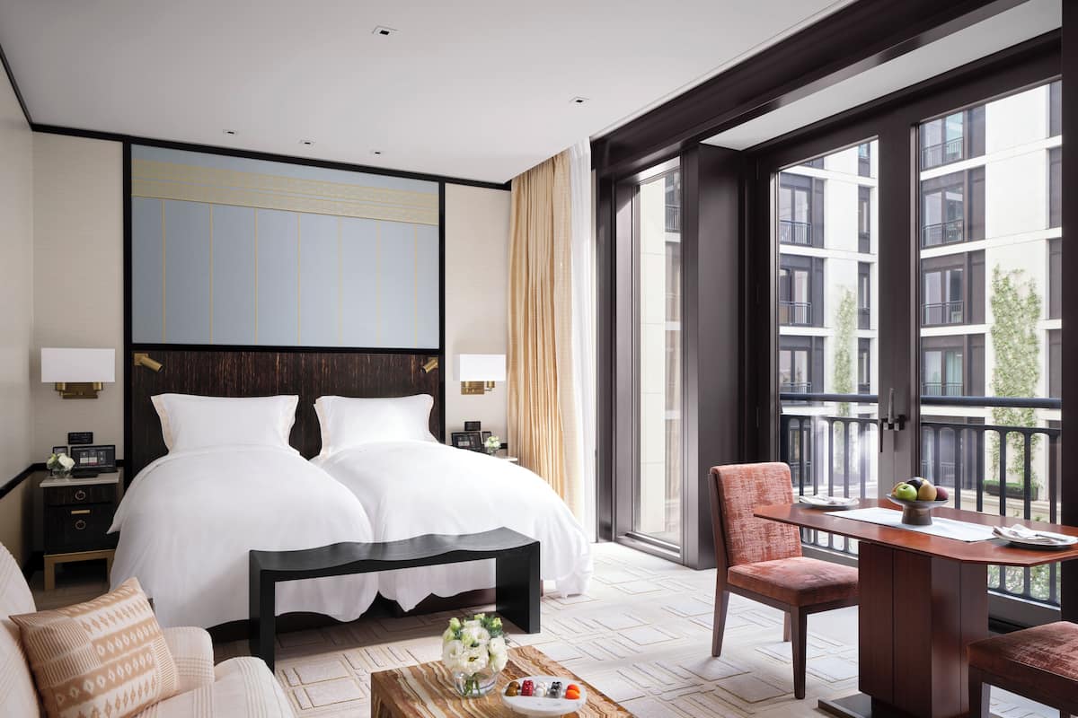 A hotel room with a large bed, a desk with a chair, a small table with a vase of flowers, and a view of the city through large windows.