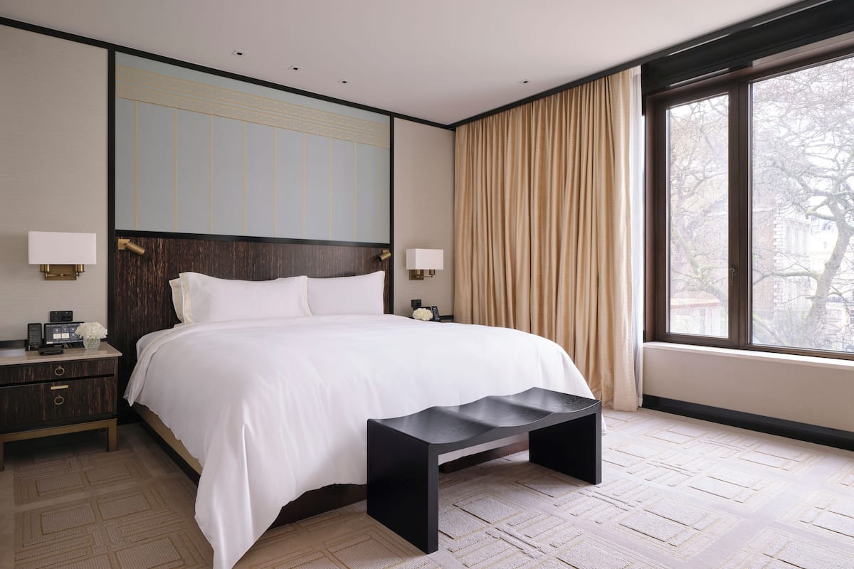 A modern hotel room with a large bed, bedside tables, a bench, and a view of the city through large windows.