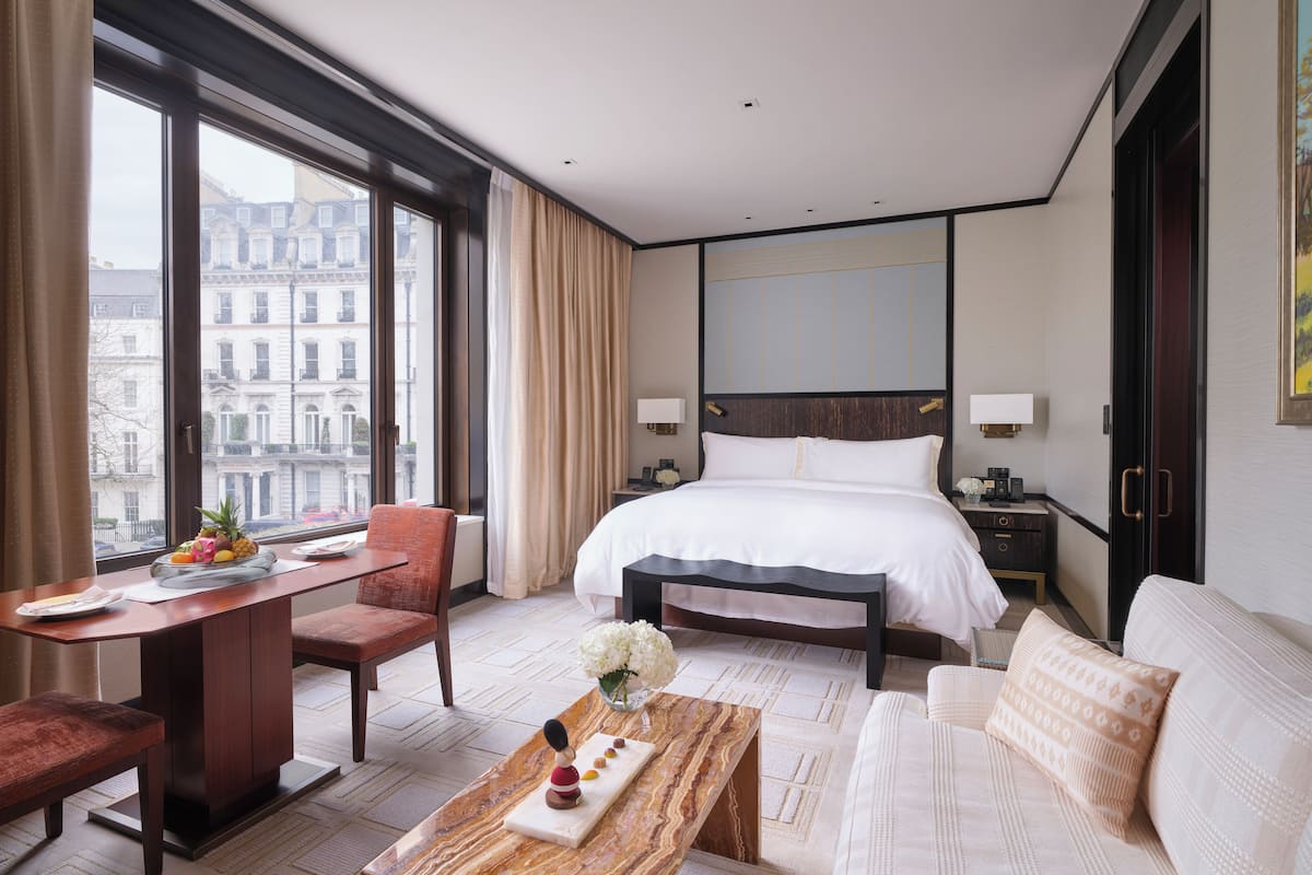 A hotel room with a large bed, a desk with a chair, a sofa, and a view of the city through large windows.