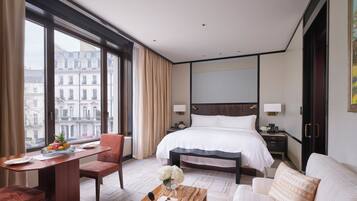 A hotel room with a large bed, a desk with a chair, a sofa, and a view of the city through large windows.