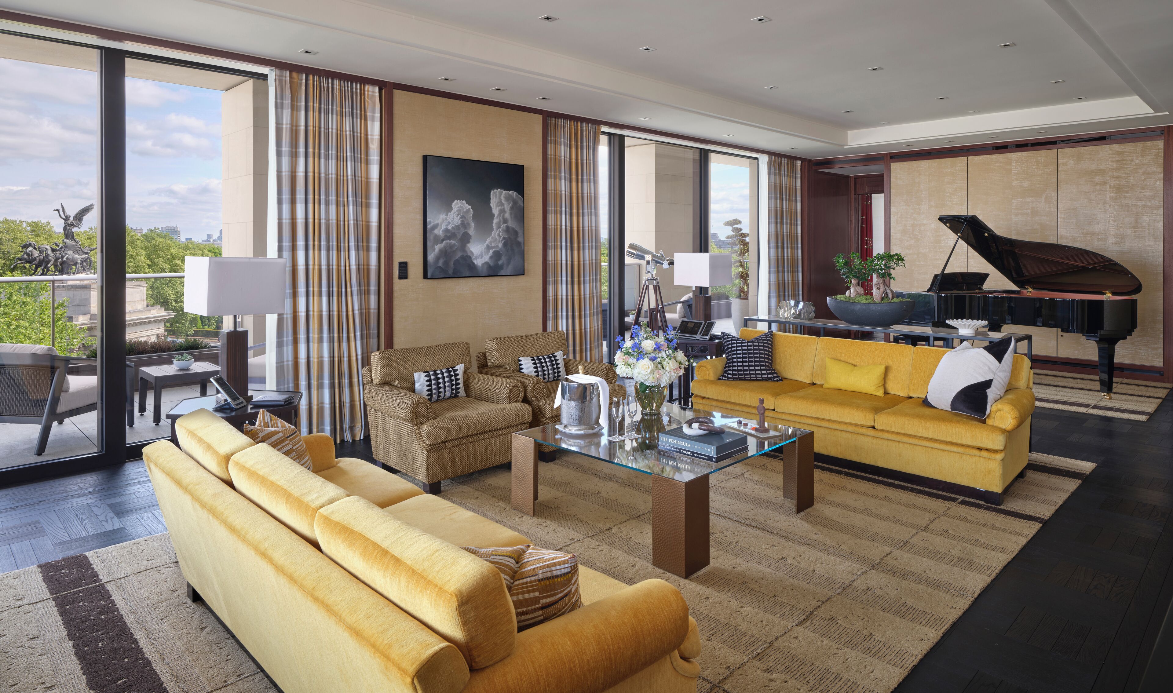 Een ruime woonkamer met een gele bank, een bruine fauteuil en een geel zitbankje. Achterin staat een piano en een beeld van een engel.