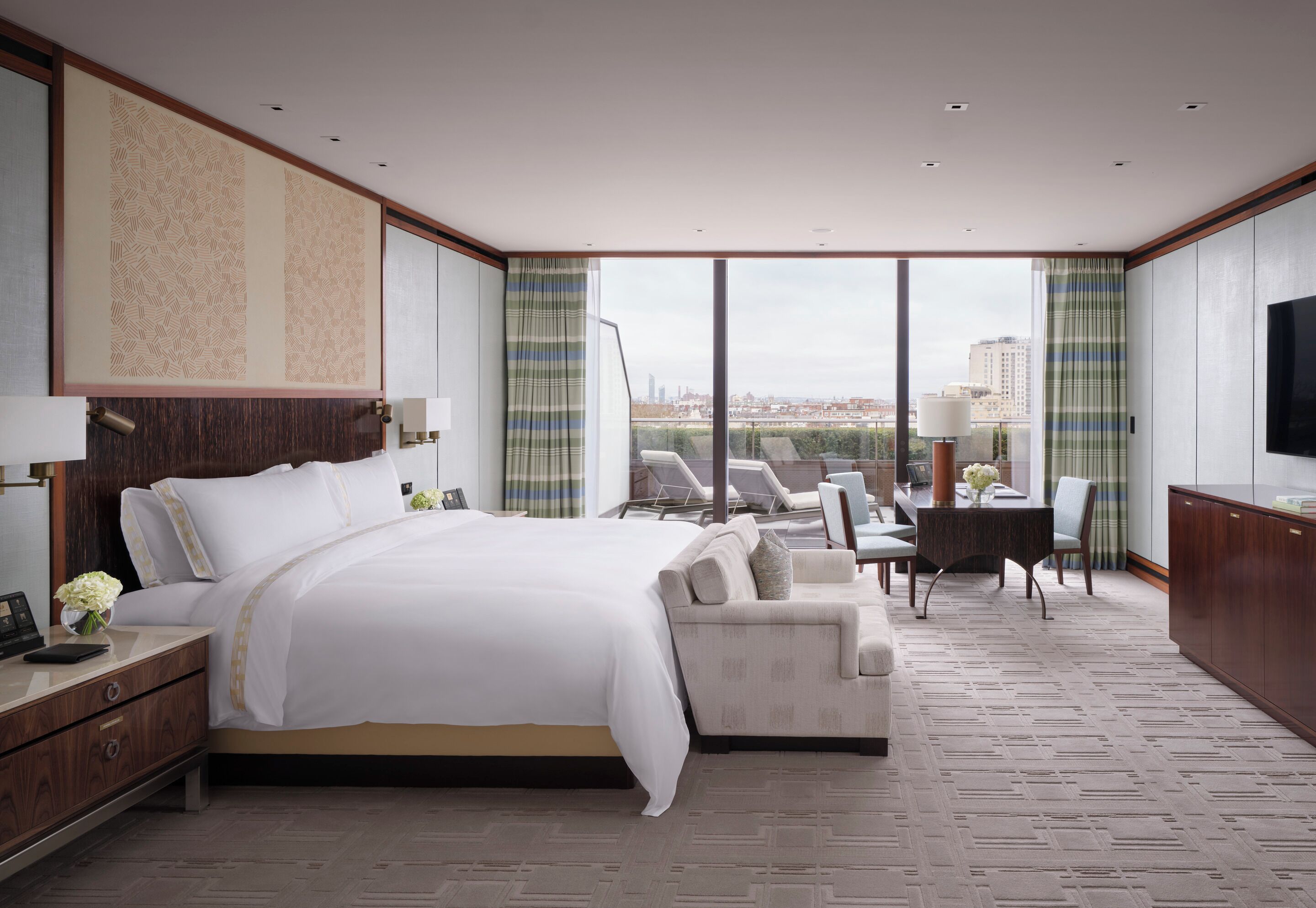 A modern hotel room with a large bed, a seating area with a table, and a city view through large windows.