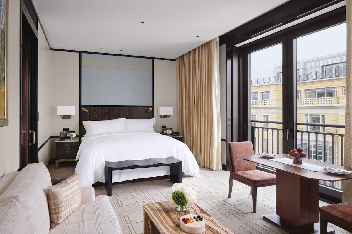 A hotel room with a large bed, a desk, a chair, a small table with a vase, and a view of buildings through large windows.