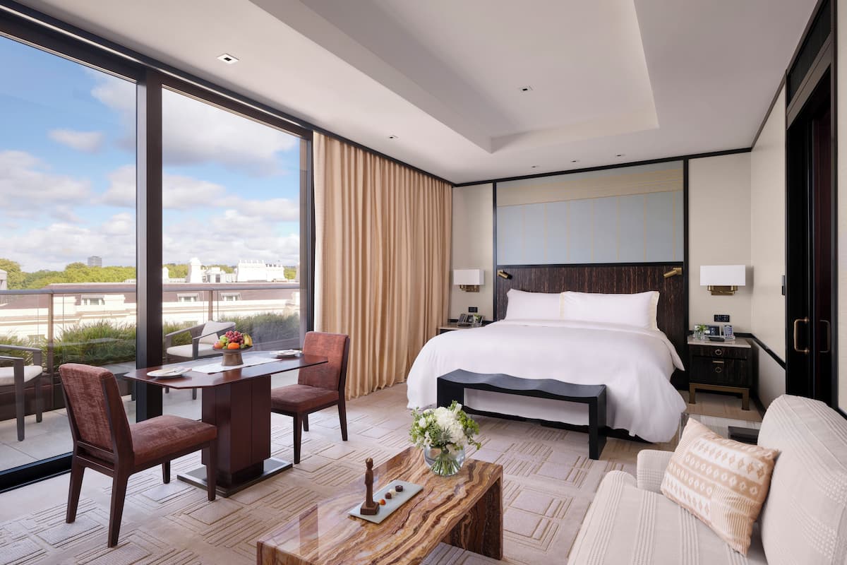 A modern hotel room with a large bed, a desk with chairs, a coffee table, and a view of the city.