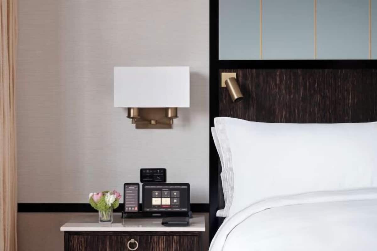 A hotel room with a bed, a bedside table with a lamp, a digital device, and a vase with flowers.