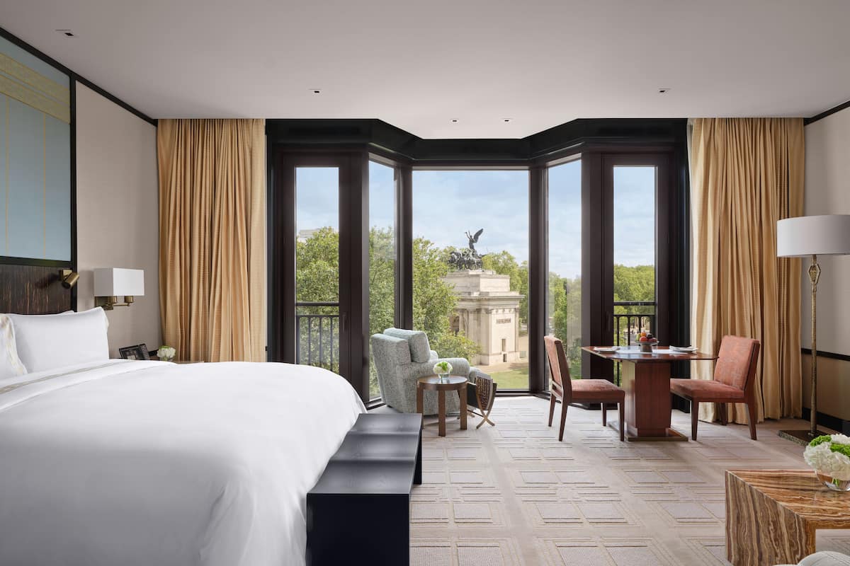 A hotel room with a large bed, a desk with a chair, a small table with a vase of flowers, and a view of a building and trees through large windows.