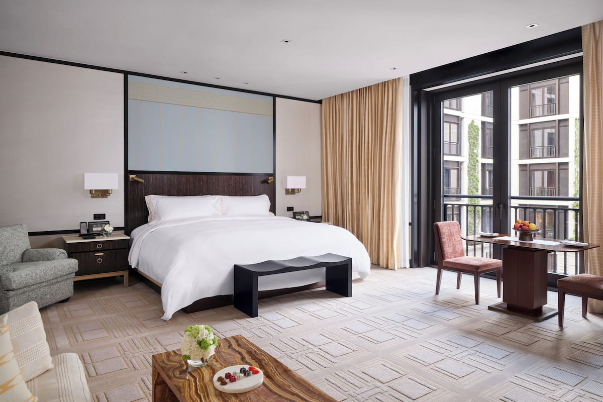A hotel room with a large bed, a desk, a chair, a small table with fruit, and a view of buildings through a large window.