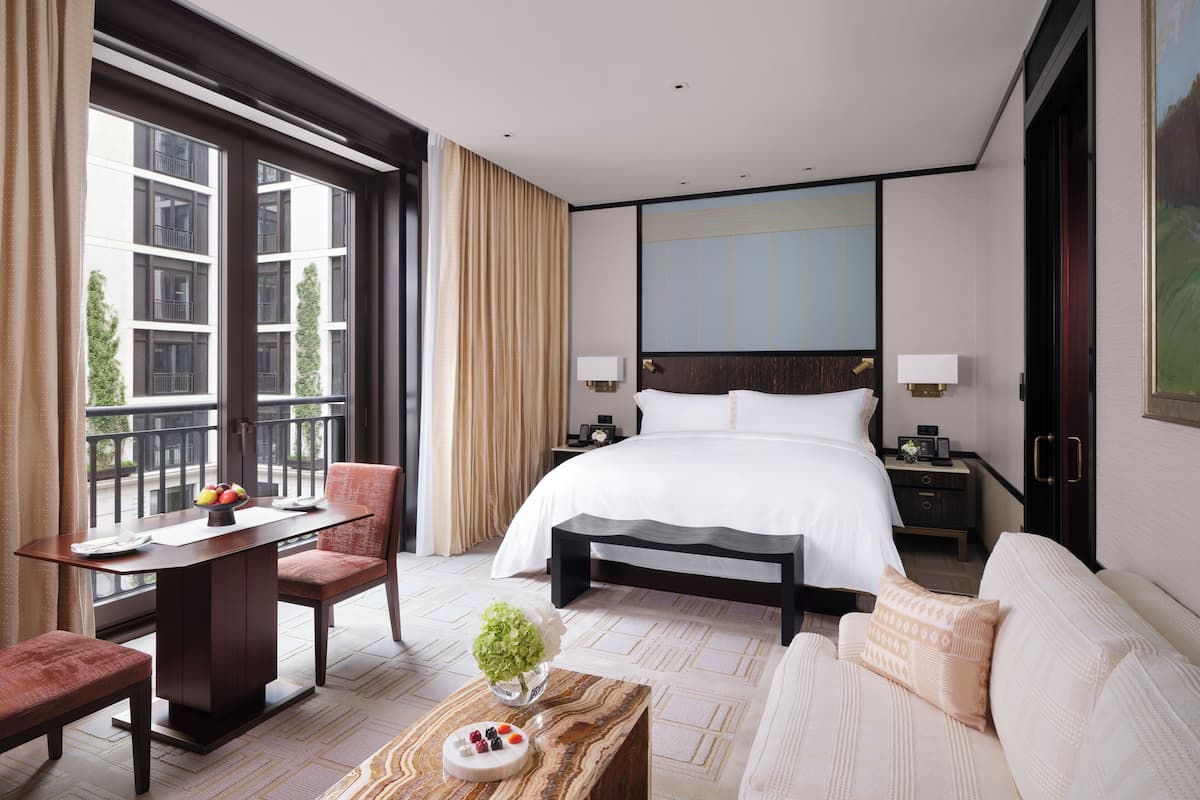 A modern hotel room with a large bed, a desk, a chair, a sofa, and a balcony with a view of the city.