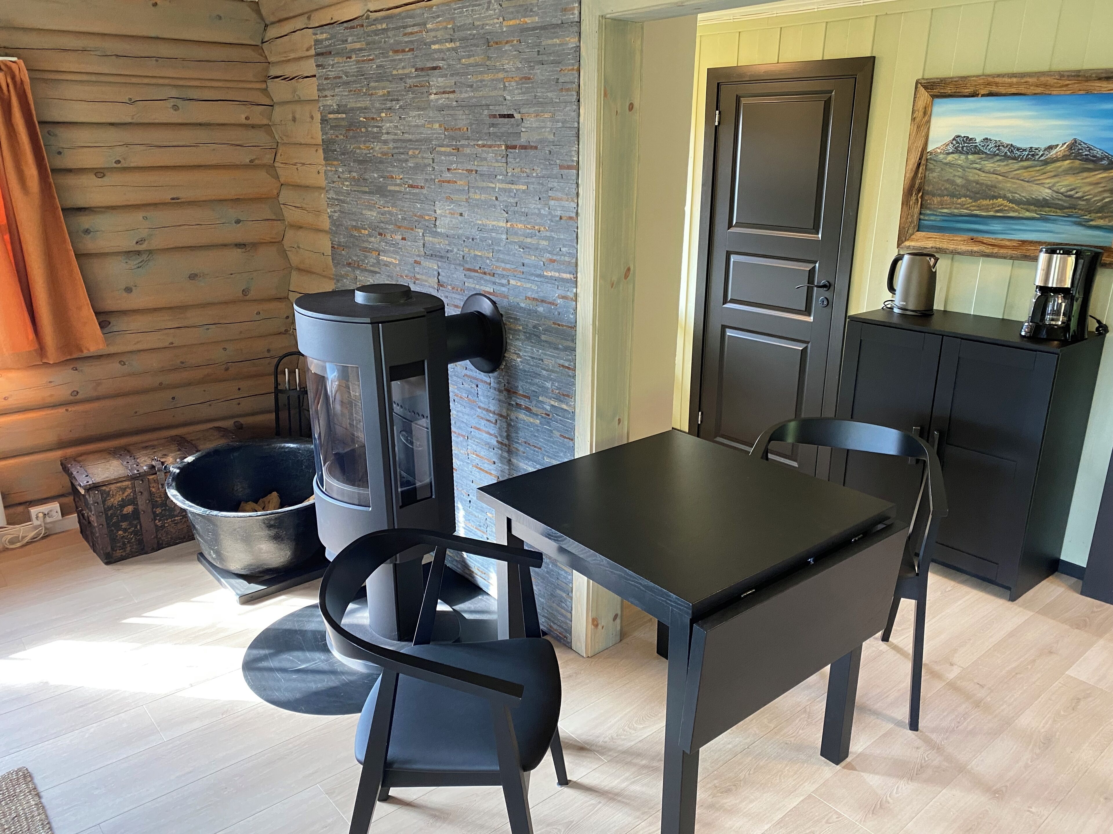A rustic cabin interior with a wooden wall, a stone wall, a black table, two black chairs, a black cabinet with a painting, and a coffee maker.