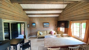 A wooden cabin interior with a dining table, chairs, a sofa, and a TV.
