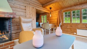 A cozy log cabin interior with a fireplace, leather armchair, dining table, and large windows offering a view of the outdoors.