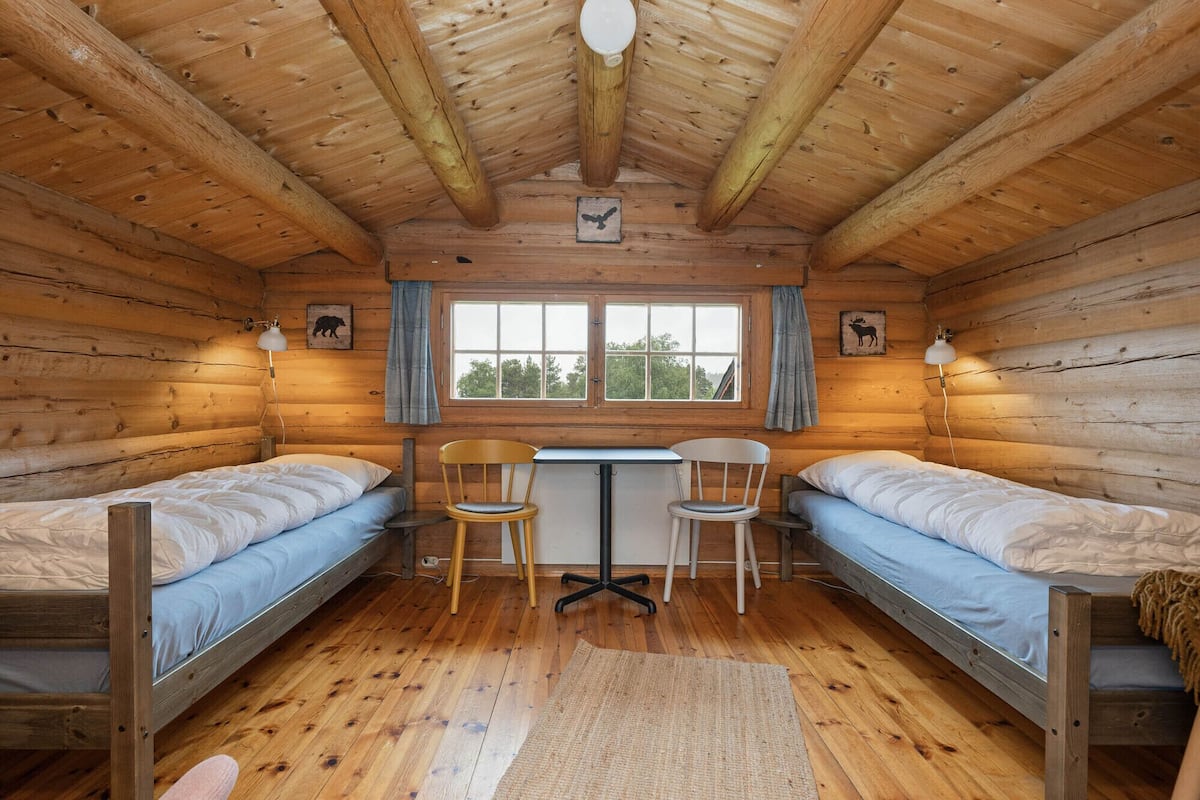 Eine Holzhütte mit zwei Etagenbetten, einem Schreibtisch, Stühlen und einem Fenster mit Vorhängen.