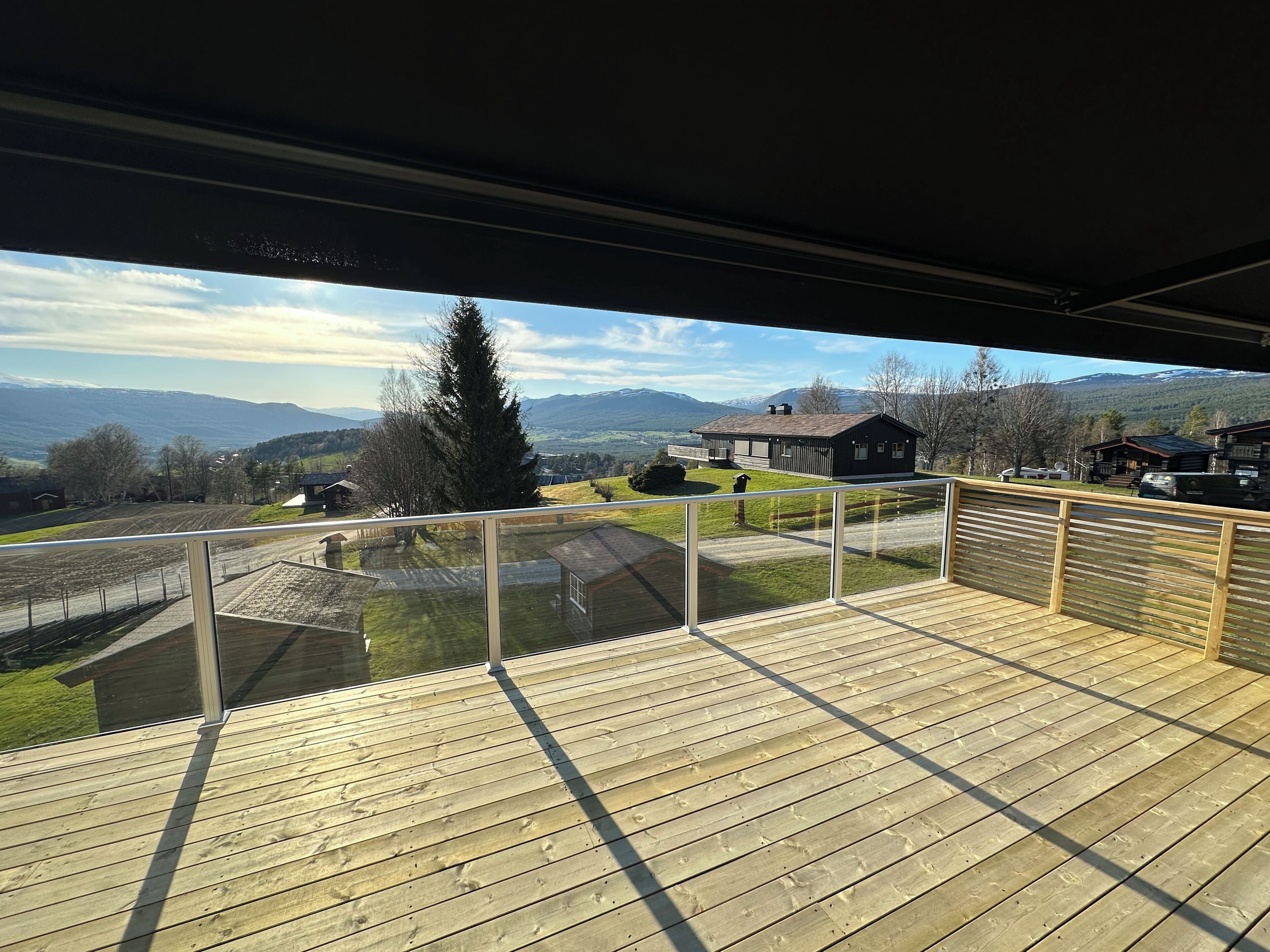 A wooden deck with a glass balustrade overlooking a rural landscape with houses and mountains.