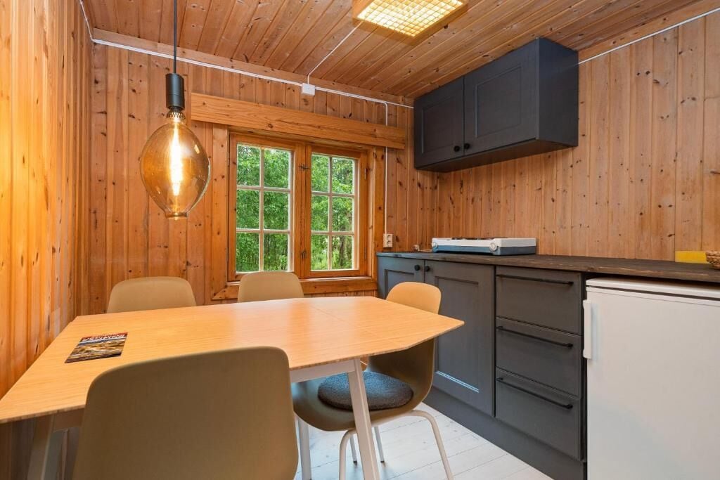A wooden interior with a dining table, chairs, a hanging light fixture, a window with a view of trees, and a kitchen area with cabinets and a refrigerator.