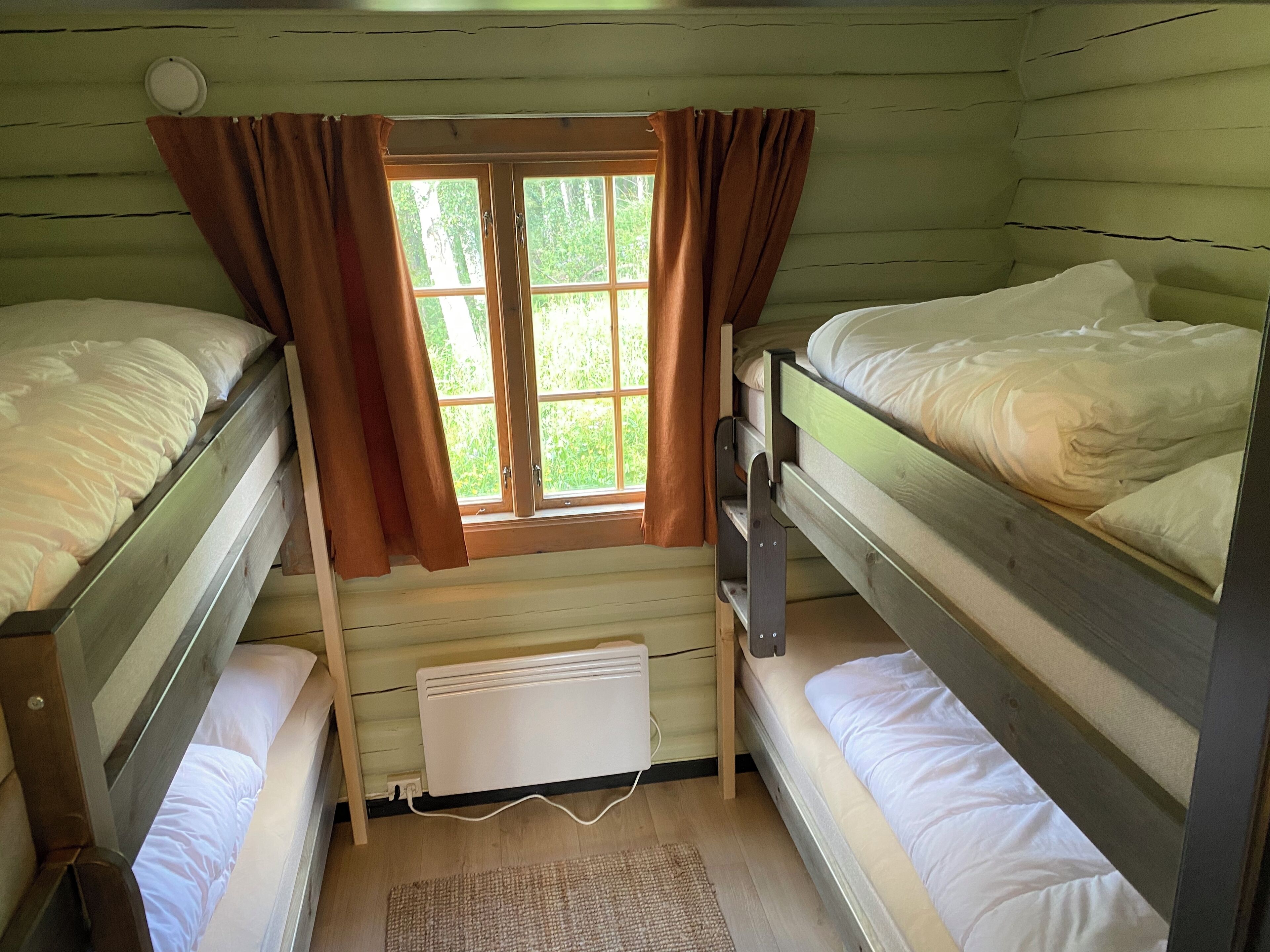 A small, cozy room with two bunk beds, a window with curtains, a radiator, and a woven mat on the floor.