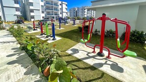 Ulkoilma-alue, jossa on kuntoiluvälineitä, parkkipaikka ja taustalla moderneja rakennuksia.