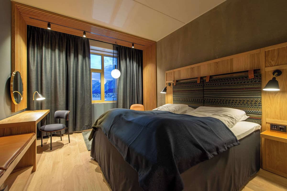 A modern bedroom with a large bed, a desk, and a chair. There is a window with curtains, a lamp, and a view of mountains.