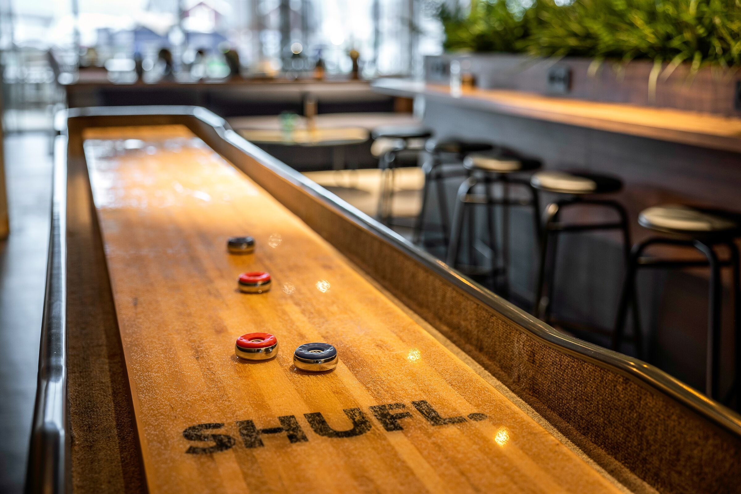 A shuffleboard table with the word "SHUFL-" visible, surrounded by bar stools and a bar area.