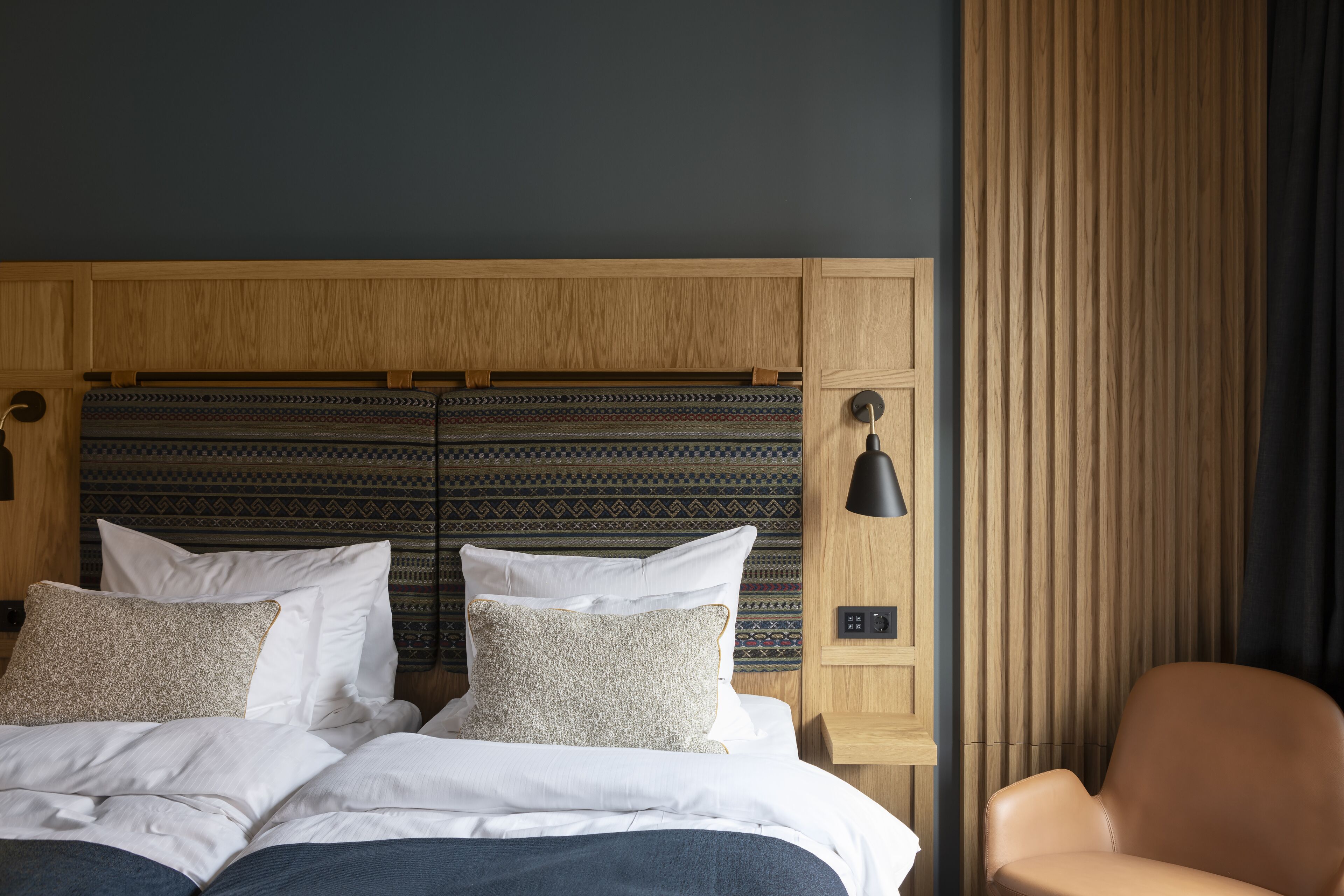 A neatly made bed with white linens and patterned pillows, a wooden headboard, and a single brown leather chair.