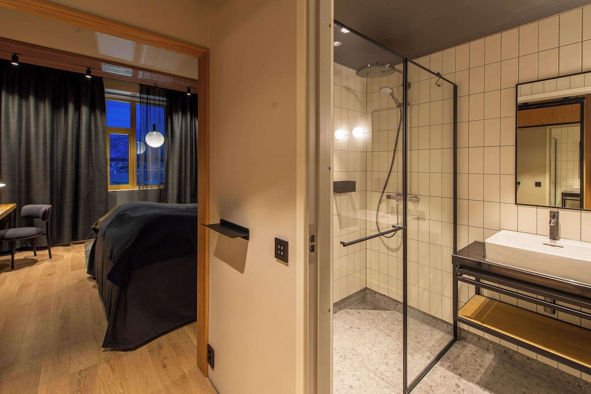 A hotel room with a bed, a desk with a chair, a window with curtains, and a shower area with a glass door.