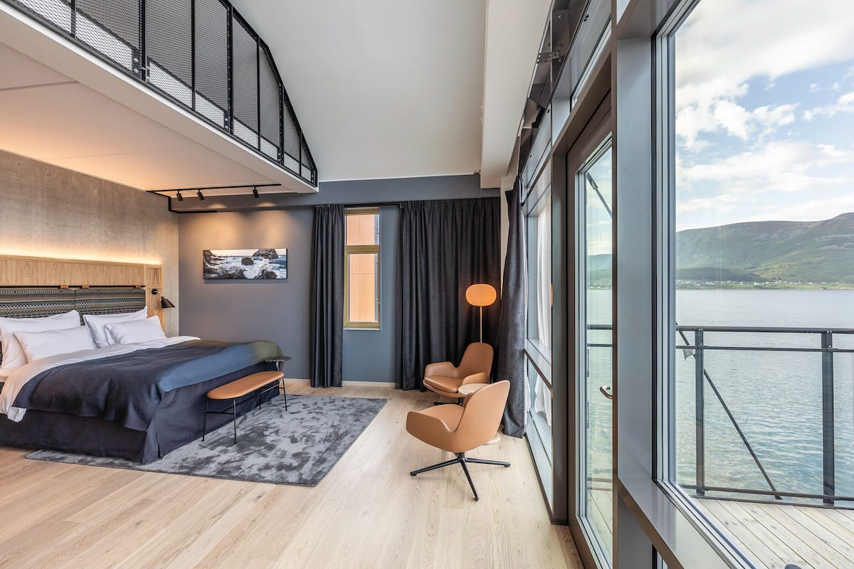 A modern bedroom with a bed, a desk, and a chair, a large window with a view of the water, and a balcony with a railing.