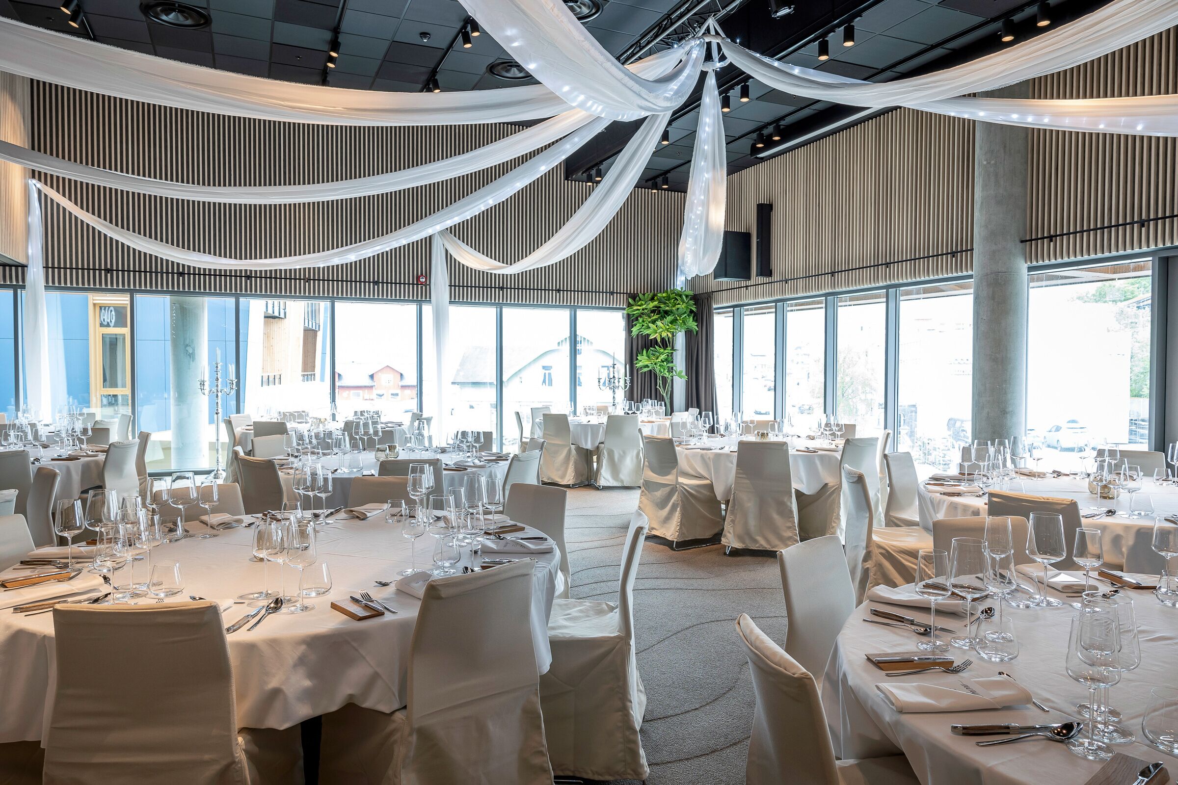 A banquet hall with round tables set for dining, large windows, and draped white fabric on the ceiling.