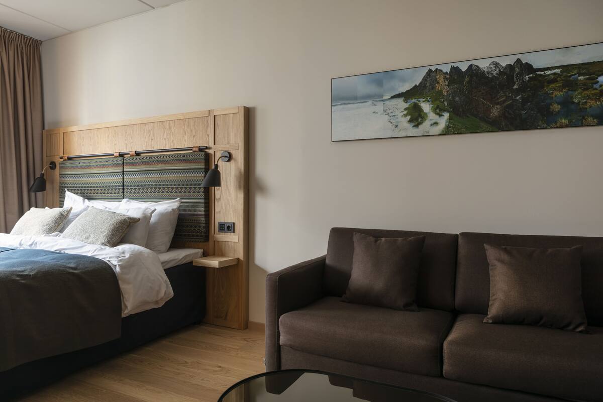 A modern hotel room with a bed, a sofa, a wooden headboard, a wall-mounted lamp, and a framed landscape painting.