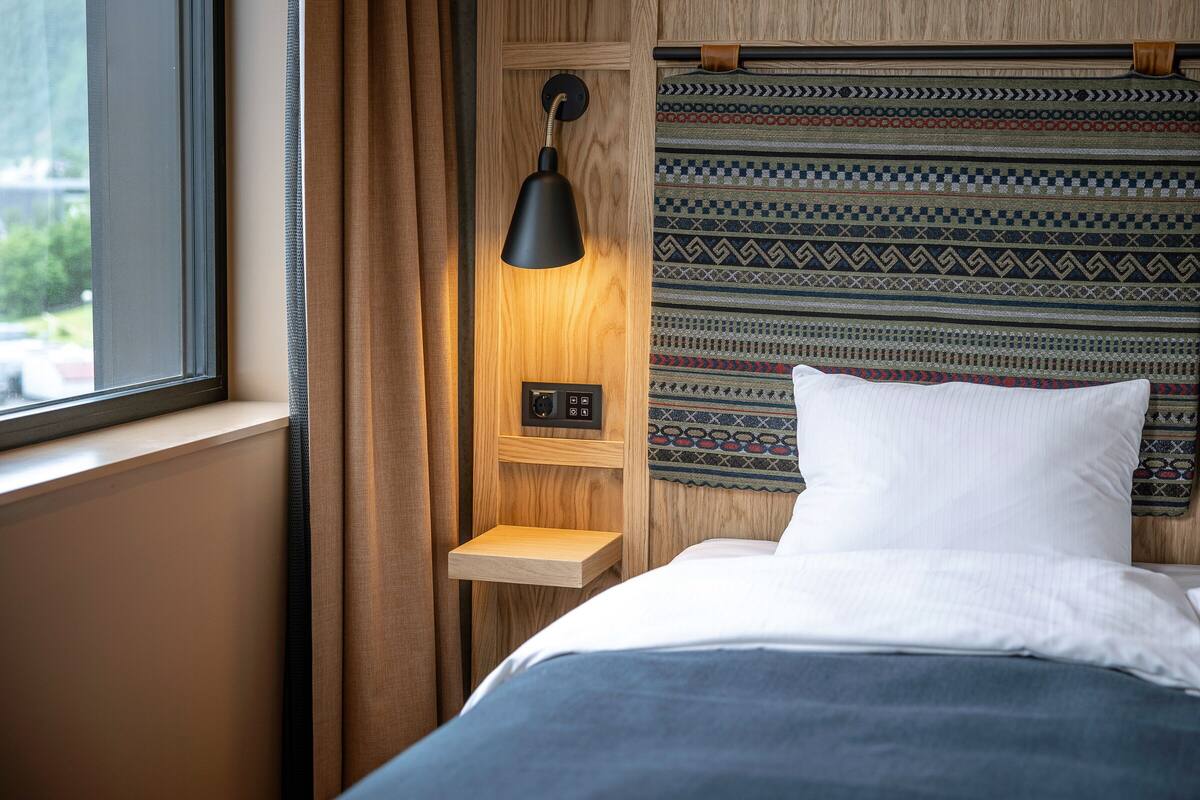A bedroom with a bed, a bedside table, a lamp, a power outlet, and a window.