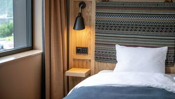 A bedroom with a bed, a bedside table, a lamp, a power outlet, and a window.