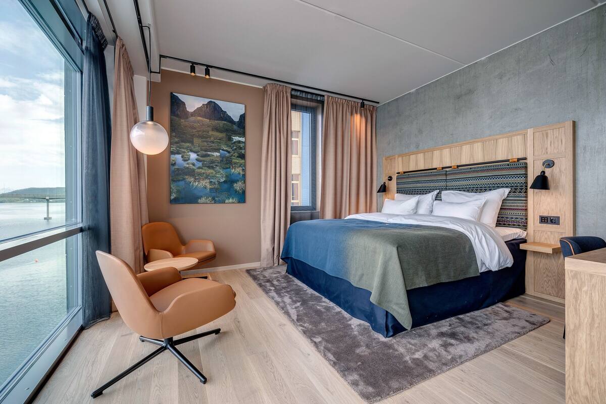 A modern hotel room with a large bed, a desk, a chair, and a view of the sea.