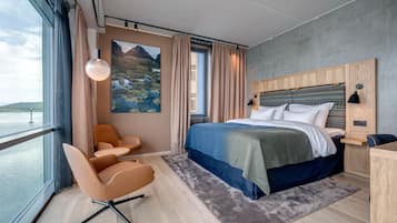 A modern hotel room with a large bed, a desk, a chair, and a view of the sea.