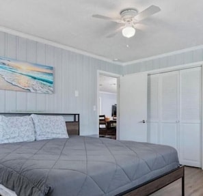 Un dormitorio con cama, ventilador de techo, un cuadro y una puerta que da a otra habitación.