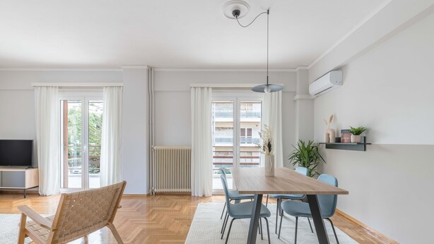 A modern living room with a wooden dining table, blue chairs, a woven chair, a TV, and a large window with curtains. - Homey 2BR Apartment in Cholargos (Papagou-Holargos)