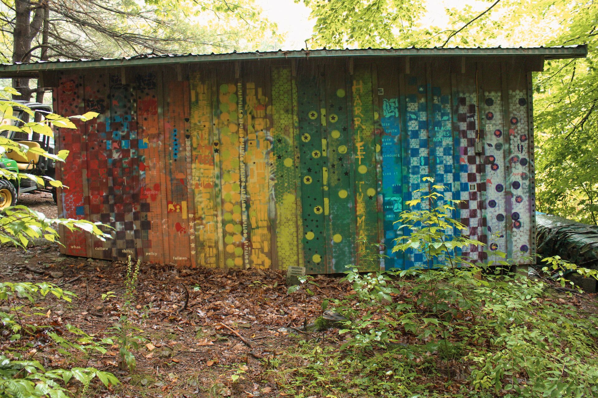 A colorful, weathered shed with graffiti and stickers, surrounded by trees and foliage.