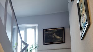 Eine Treppe mit Holzstufen und weißen Geländern, ein grauer Stuhl mit weißem Kissen, ein gerahmtes Bild an der Wand und ein Blumentopf vor dem Fenster.