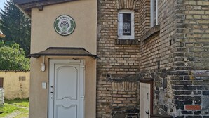 Ein Gebäude mit einer weißen Tür, einem kleinen Fenster mit Spitzenvorhängen und einem Schild mit einem Logo und Jahreszahl.