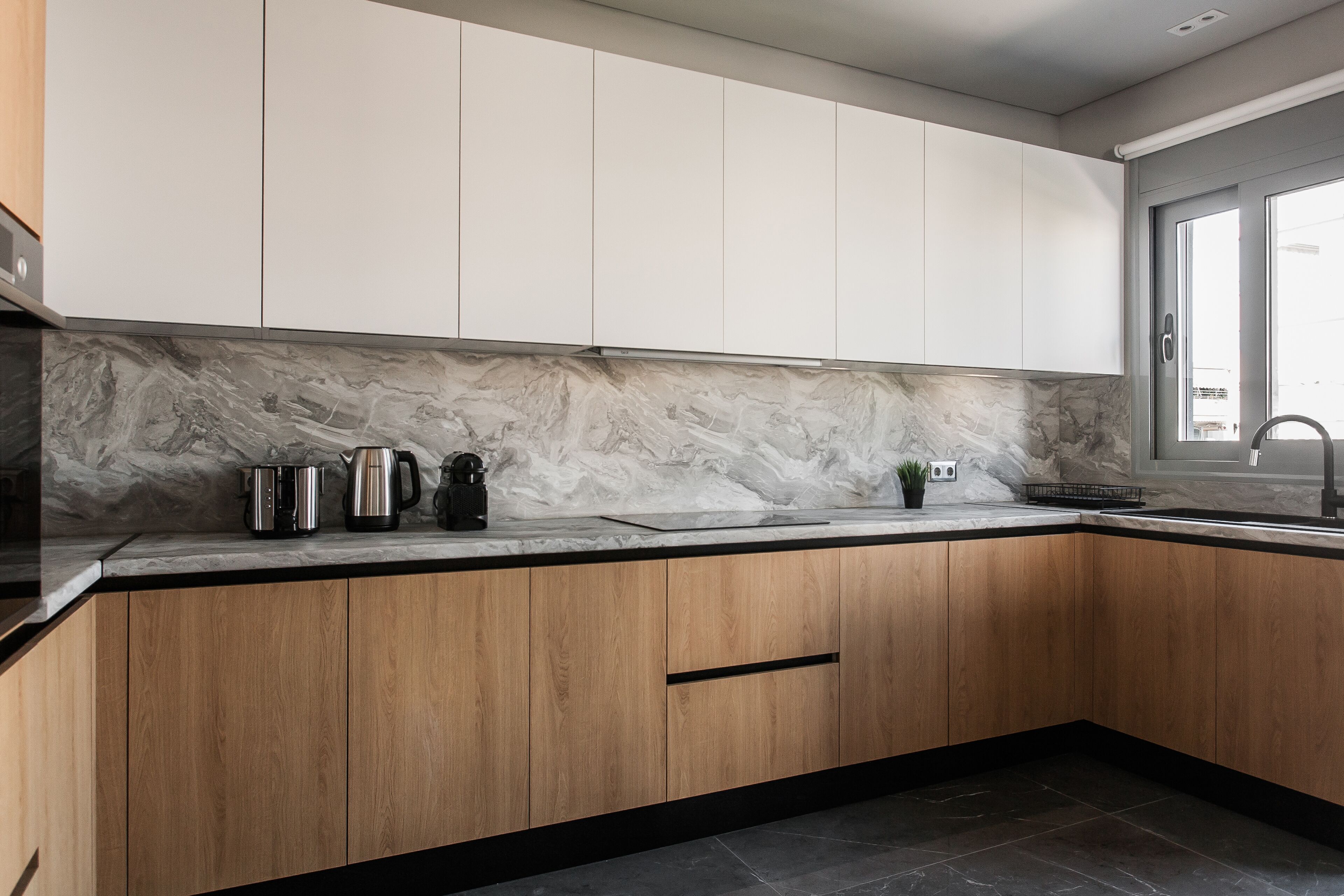 A modern kitchen with wooden cabinets, a marble backsplash, and a large window providing natural light.