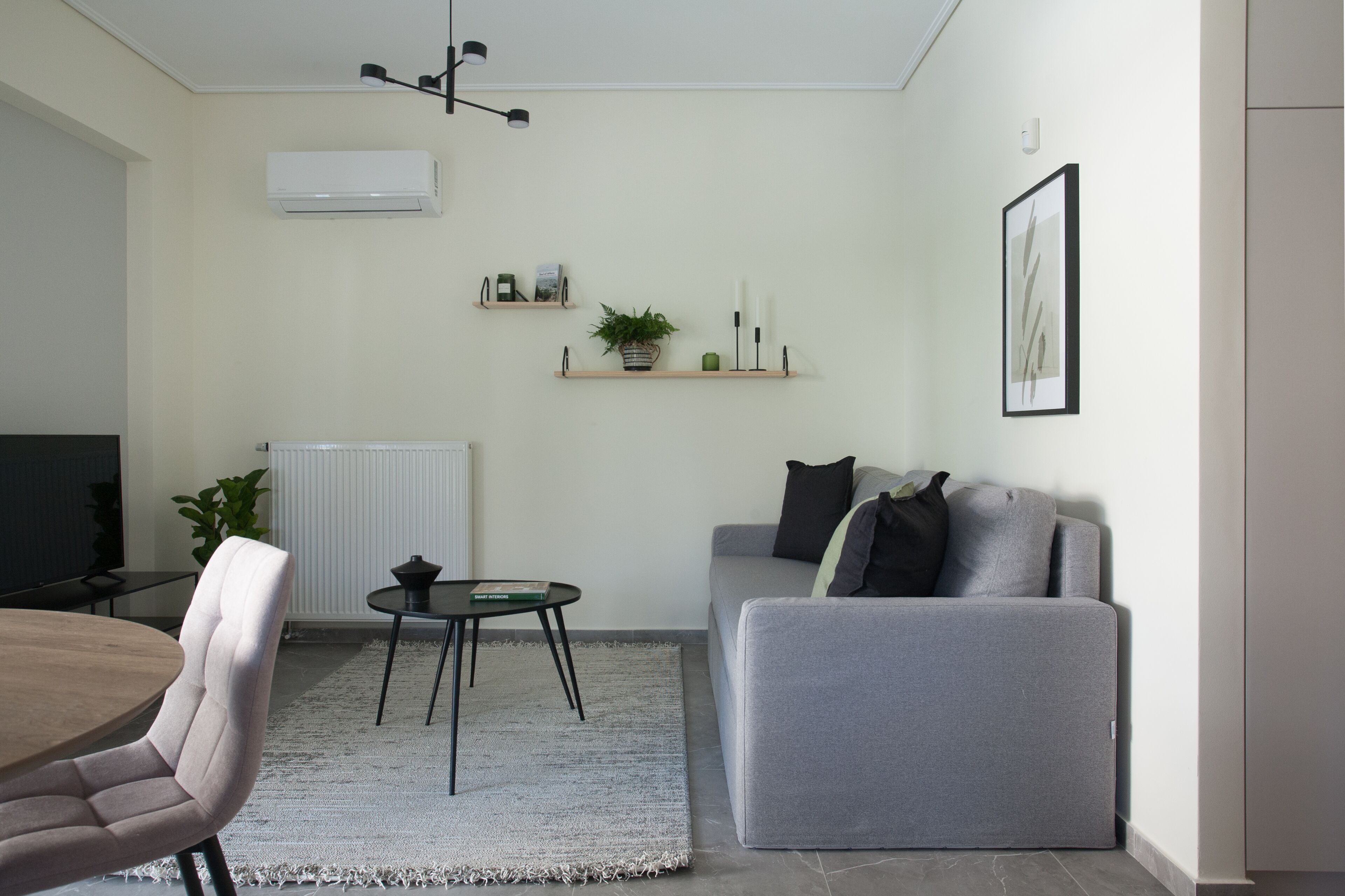 Una sala moderna con un sofá gris, una mesa redonda y un televisor. Hay dos estantes con objetos decorativos y una planta.