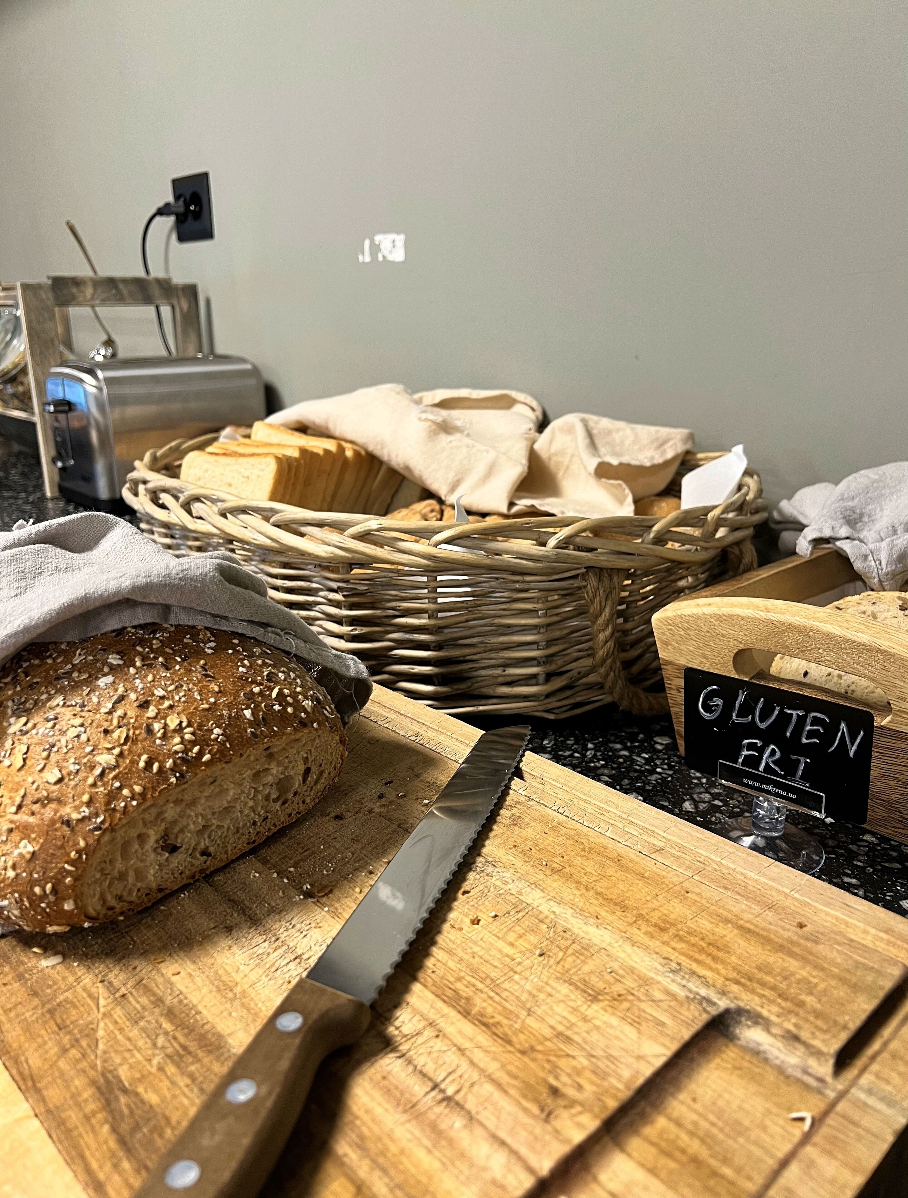 Una barra de pan sobre una tabla de cortar con un cuchillo, una cesta de pan y un cartel que dice 'GLUTEN FREE'.