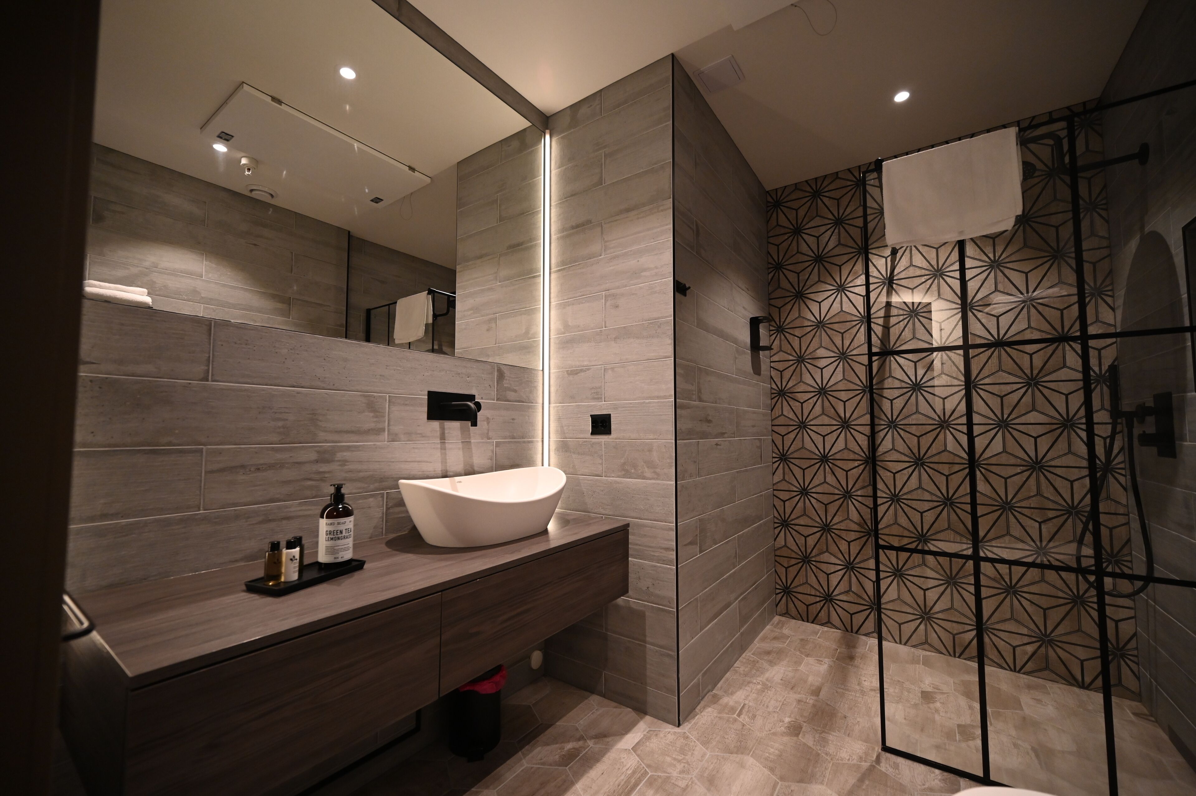 Un baño moderno con un espejo grande, un lavabo sobre encimera y un revestimiento de pared para ducha con motivos geométricos.