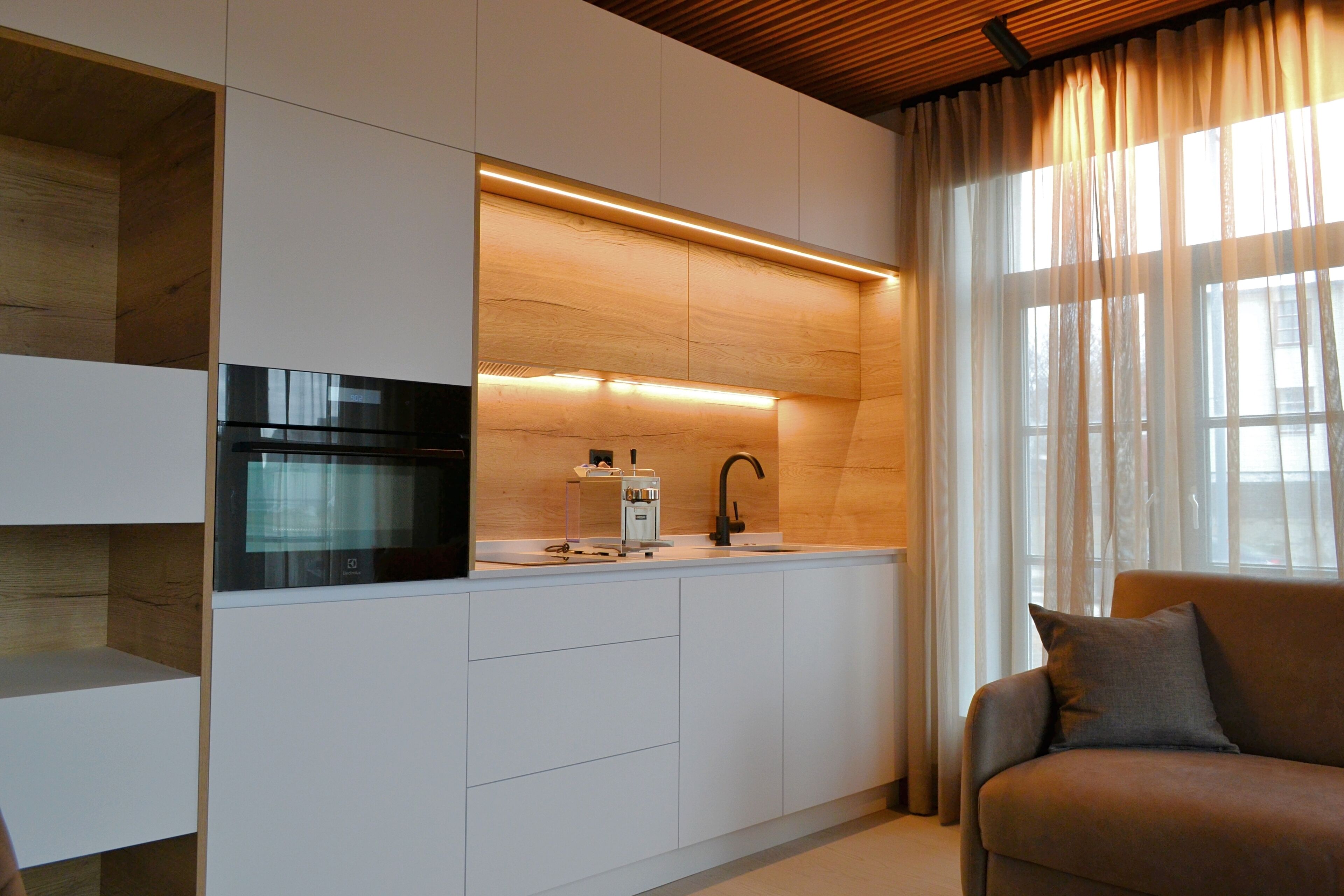 A modern kitchen with a built-in oven, a sink, and a coffee machine. There is a wooden ceiling and a window with sheer curtains.
