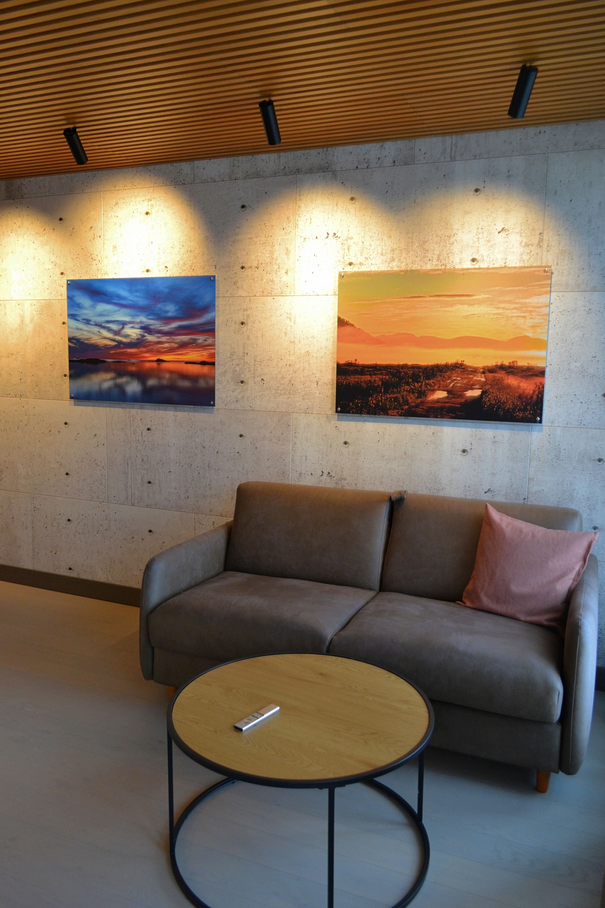 A modern lounge area with a sofa and a round coffee table, two wall-mounted paintings, and a wooden ceiling with recessed lighting.
