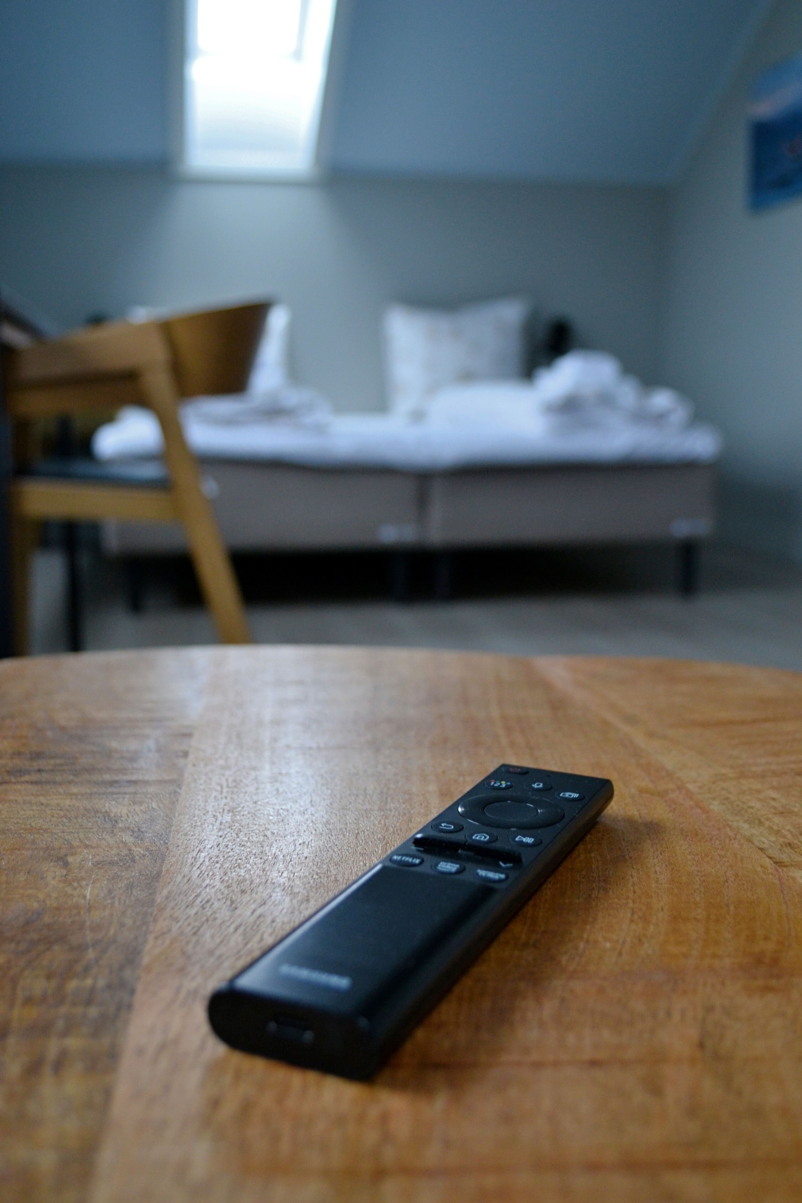 A remote control on a wooden table in a room with a bed and a chair in the background.