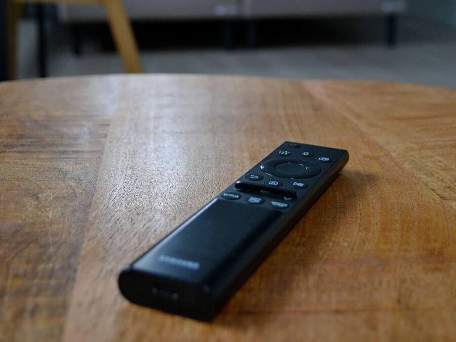 A remote control on a wooden table in a room with a bed and a chair in the background.