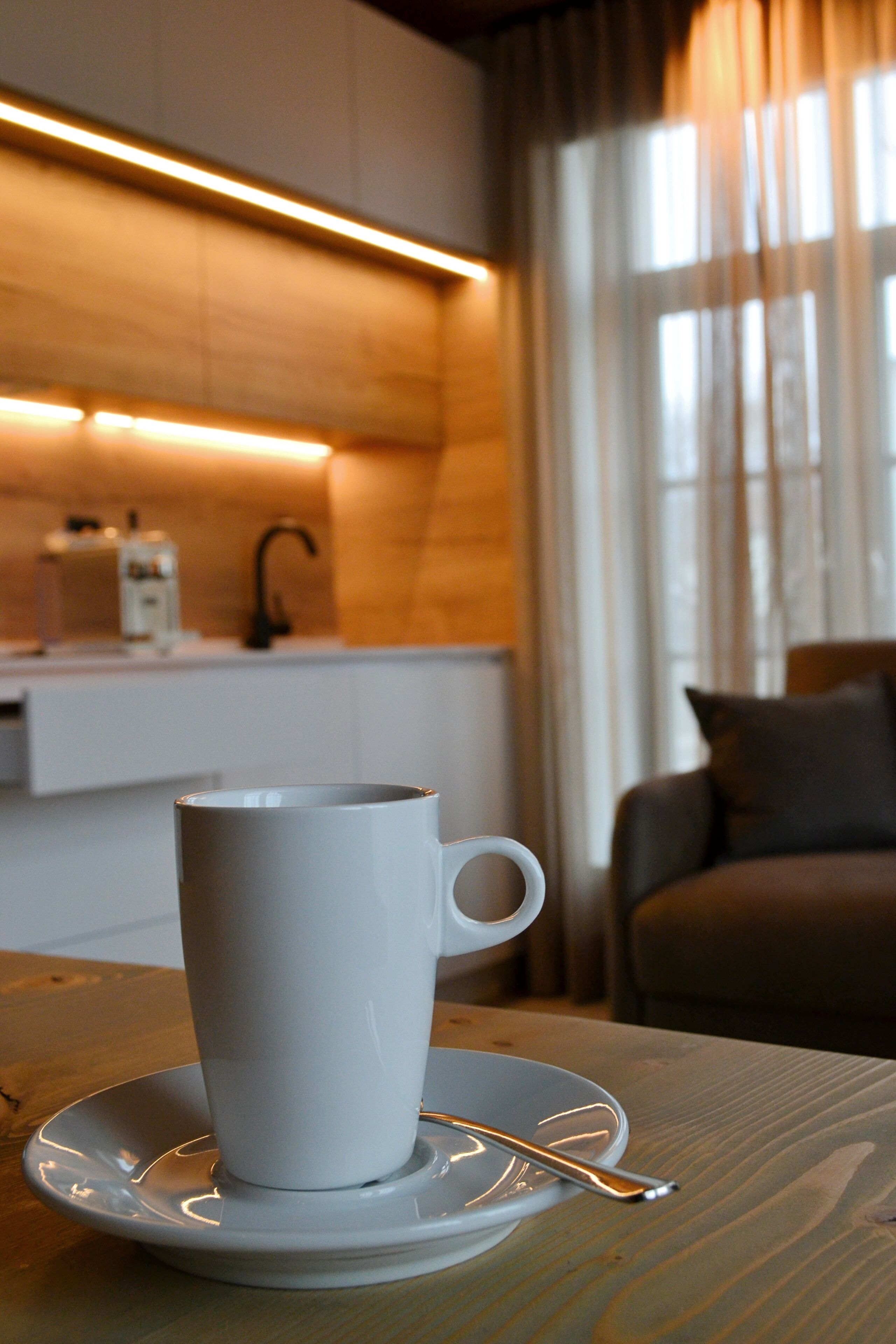 Una taza blanca sobre un plato con una cuchara en una mesa de madera. En el fondo, se observa una zona de cocina con una encimera, un lavabo y una ventana con cortinas.