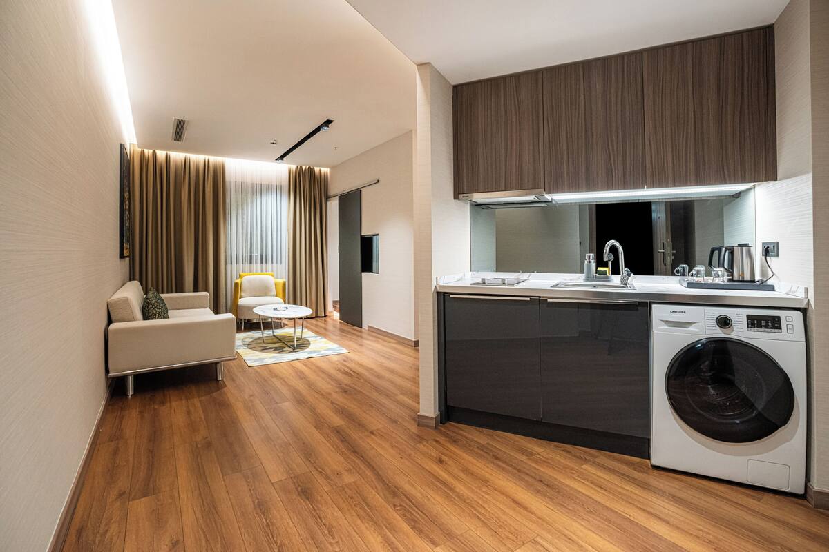 Una cocina moderna con lavadora integrada, un comedor con mesa redonda y una sala de estar con sofá y cortinas.