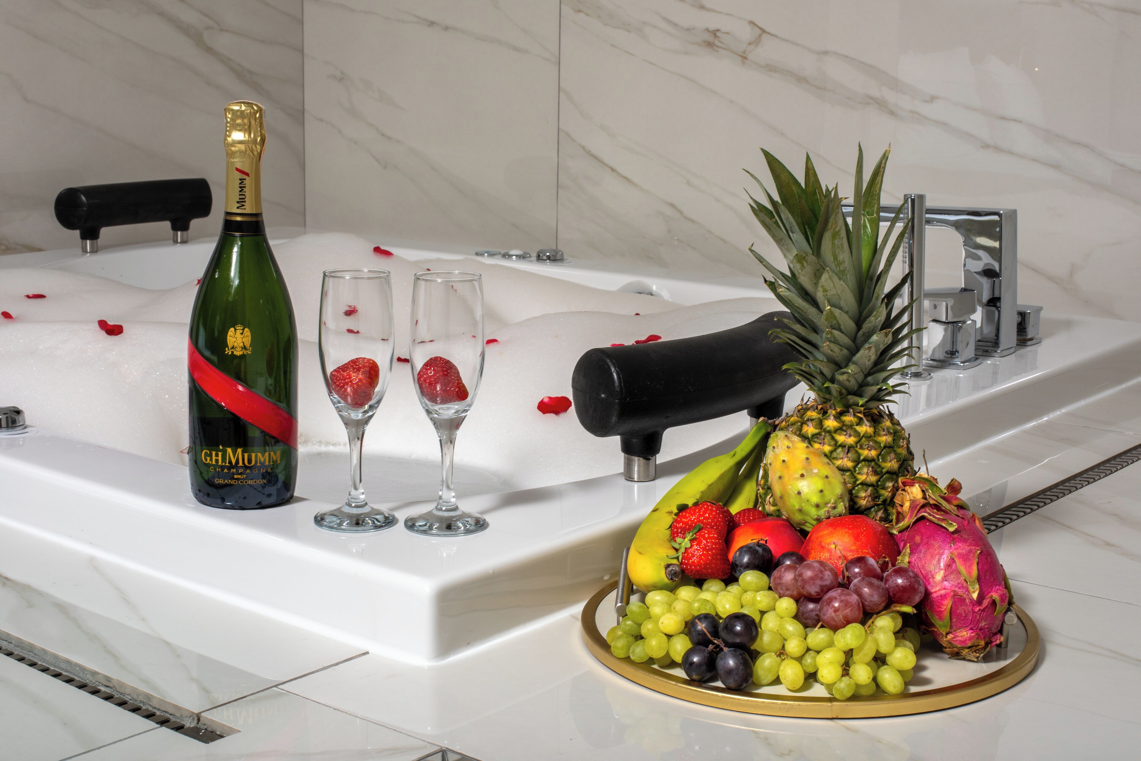 A bathroom with a bathtub, a bottle of champagne, two glasses, and a fruit platter.
